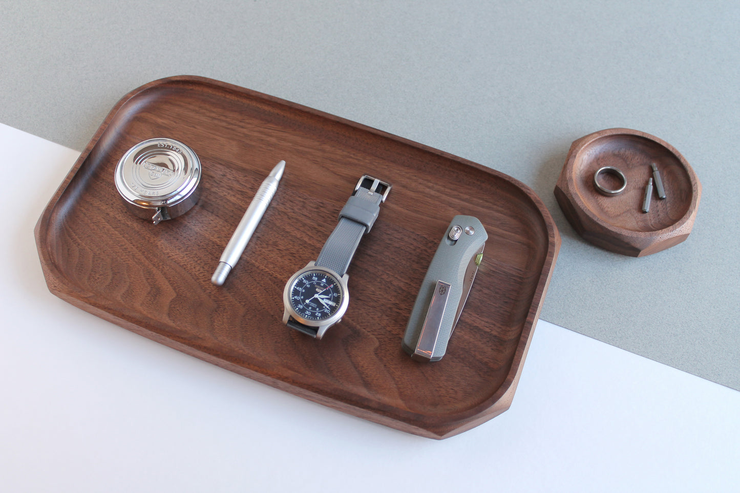 Genesis Tray and Genesis dish in Walnut wood on gray and white angled background. Angled view of tray with pocket knife, watch, pen and tape measure on tray. The dish had a ring and two Torx bit drivers.