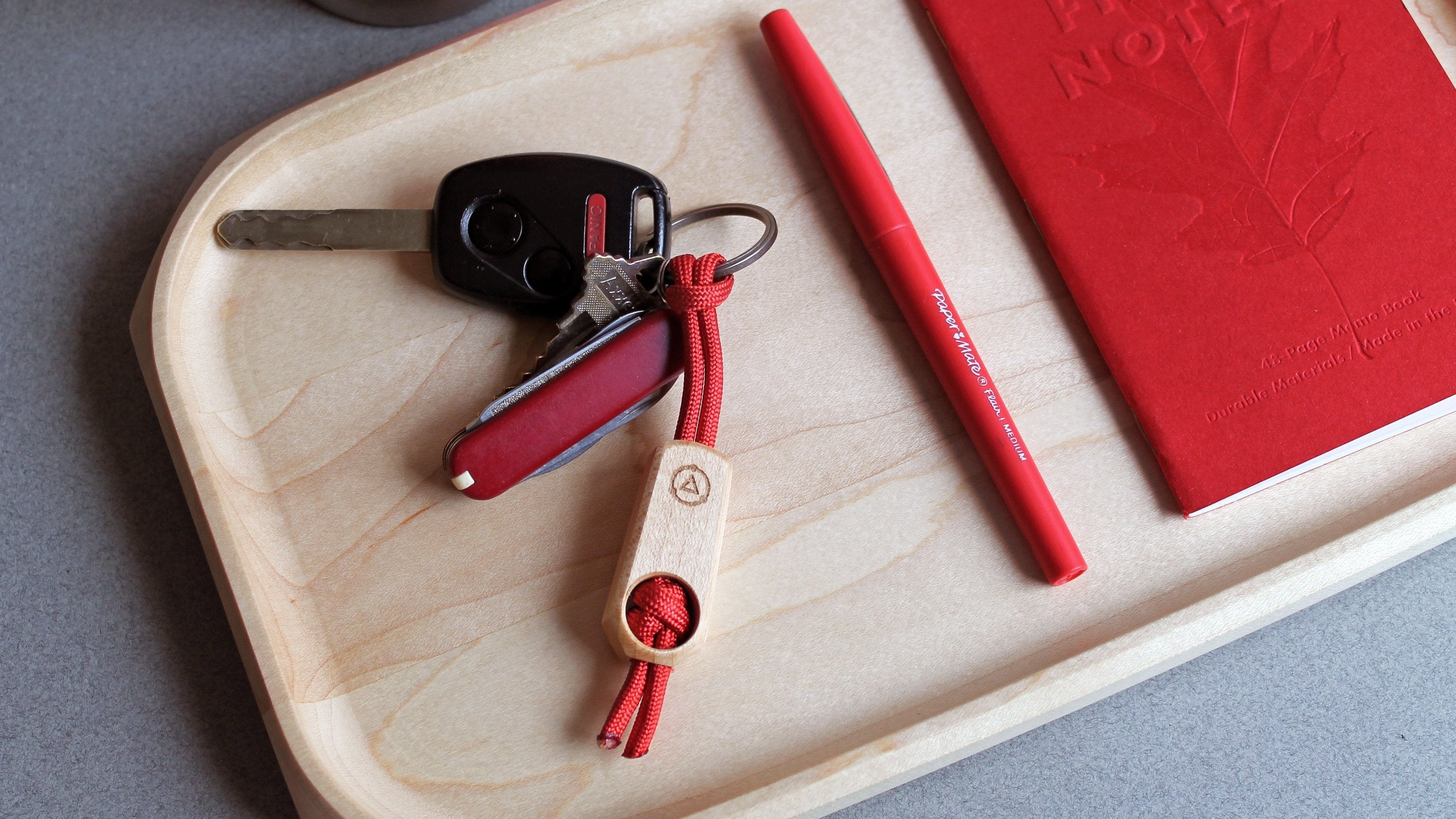 Core Key Fob with Titanium Key Ring, Paracord Lanyard and Maple Fob. Key Fob shown in red on Maple tray with red pocket knife, keys, pen and notebook.
