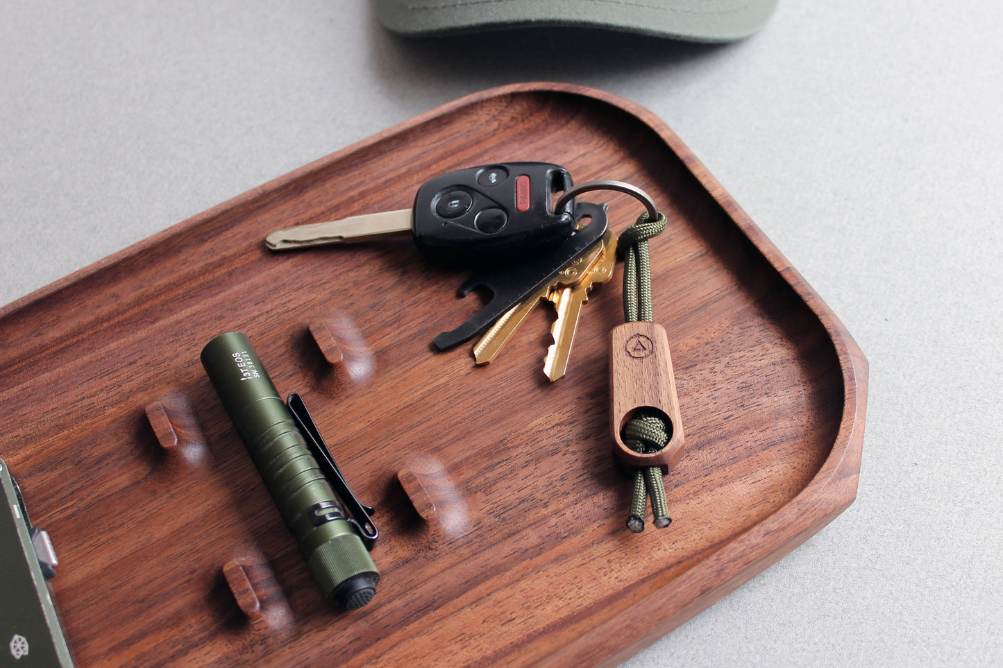 Core Key Fob with Titanium Key Ring, Paracord Lanyard and Maple Fob. Detailed view of Walnut Key Fob with olive green lanyard with keys on a walnut tray with olive green flashlight, knife and hat.